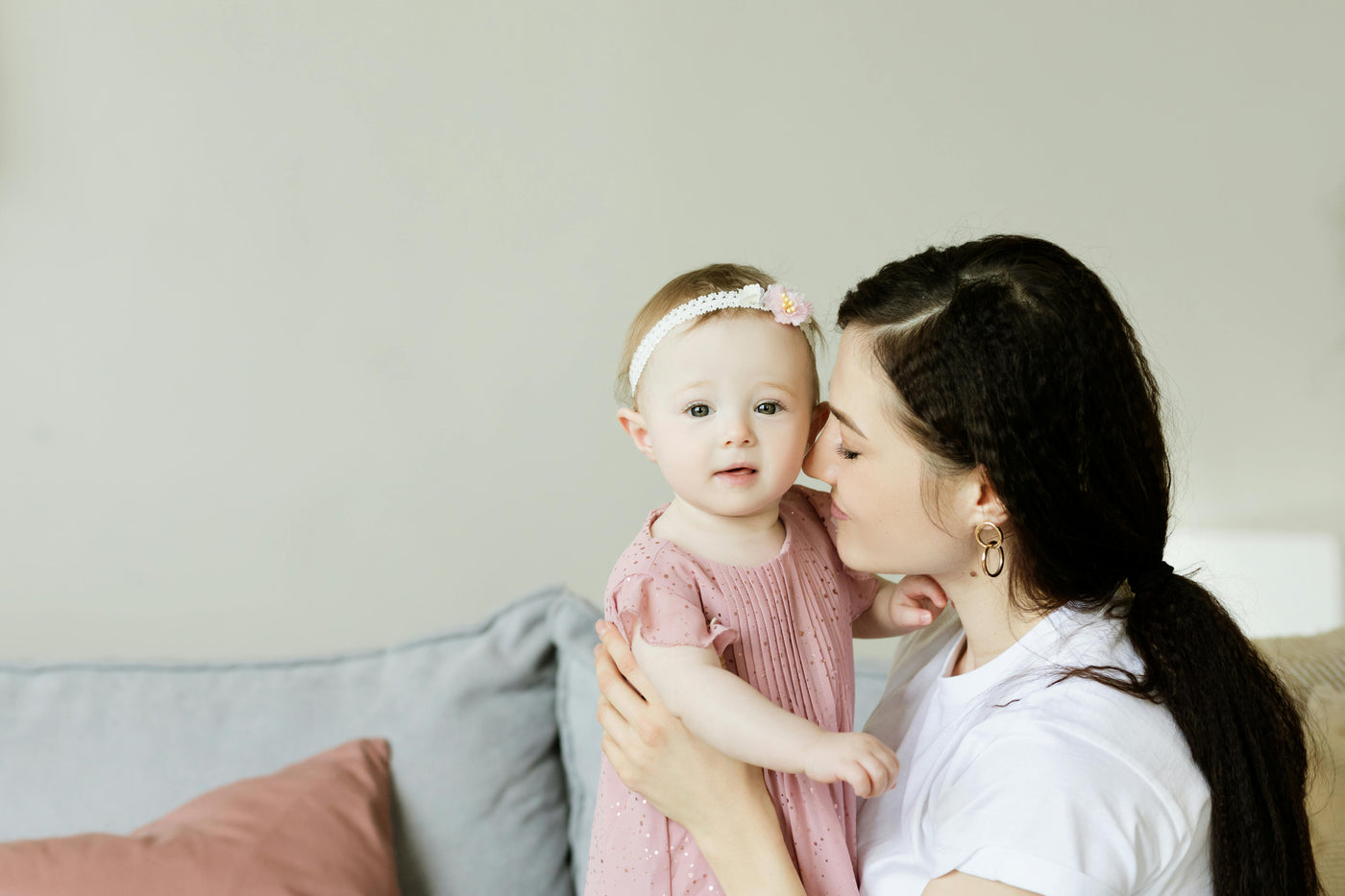 A baby in a pink dress and white headband looks at the camera. Her mother, with black hair wearing a white shirt, is facing the baby, is smiling and holding her close.