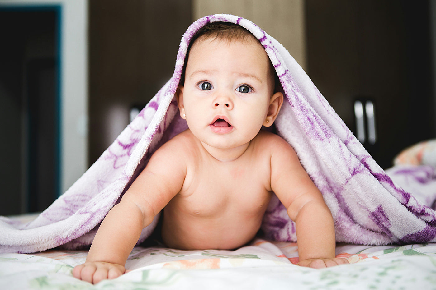 Baby looking up from under a pink blanket, looking into the camera.