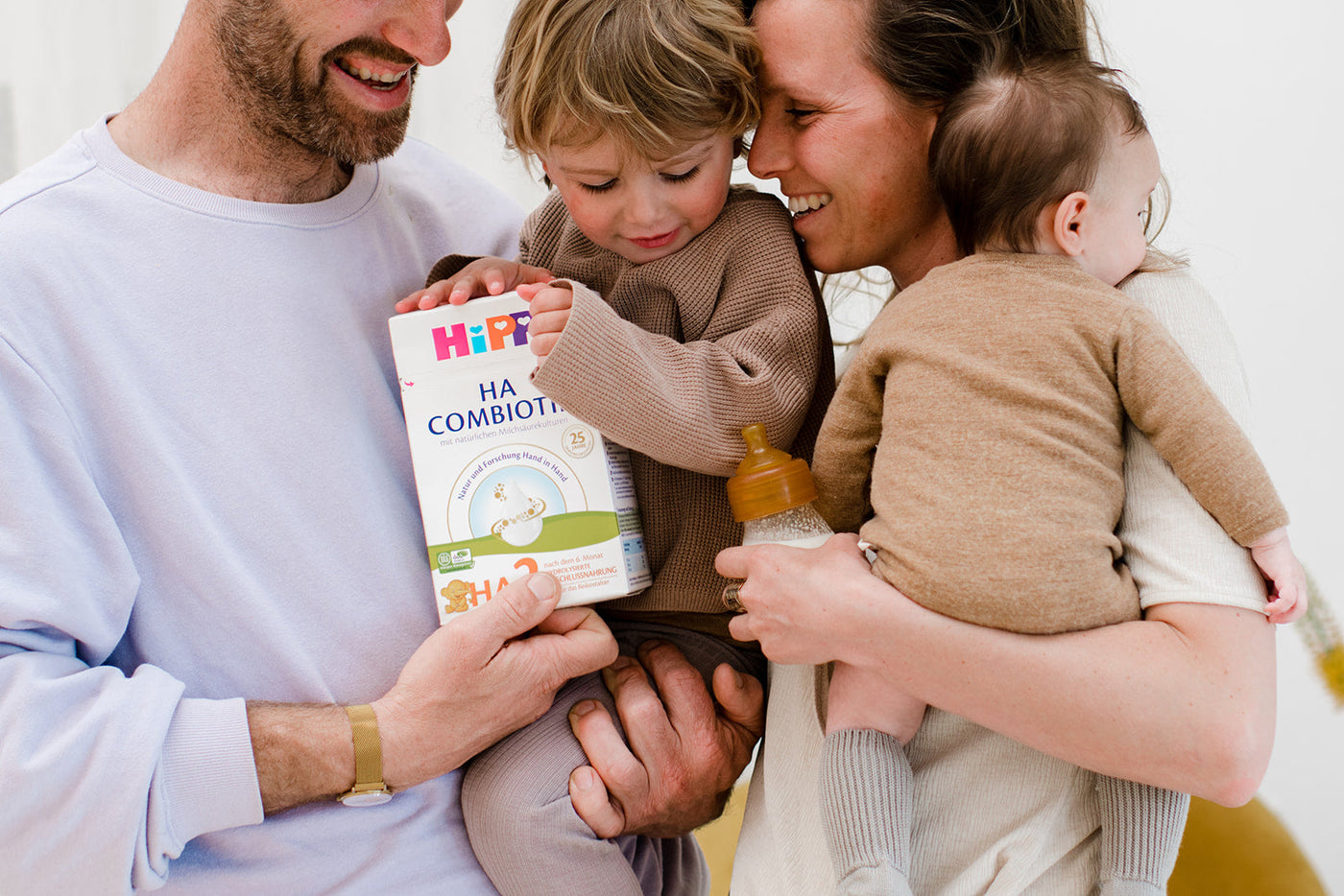 A mom and dad with their two children. The dad is holding the toddler and the mom is holding the baby. The toddler is holding a box of HiPP HA Combiotik. They are all smiling.
