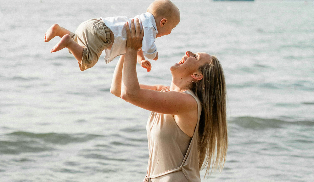 A mom and baby on the beach in summer. The mom , with long brown hair and a beige dress, is smiling and lifting the baby up into the air.