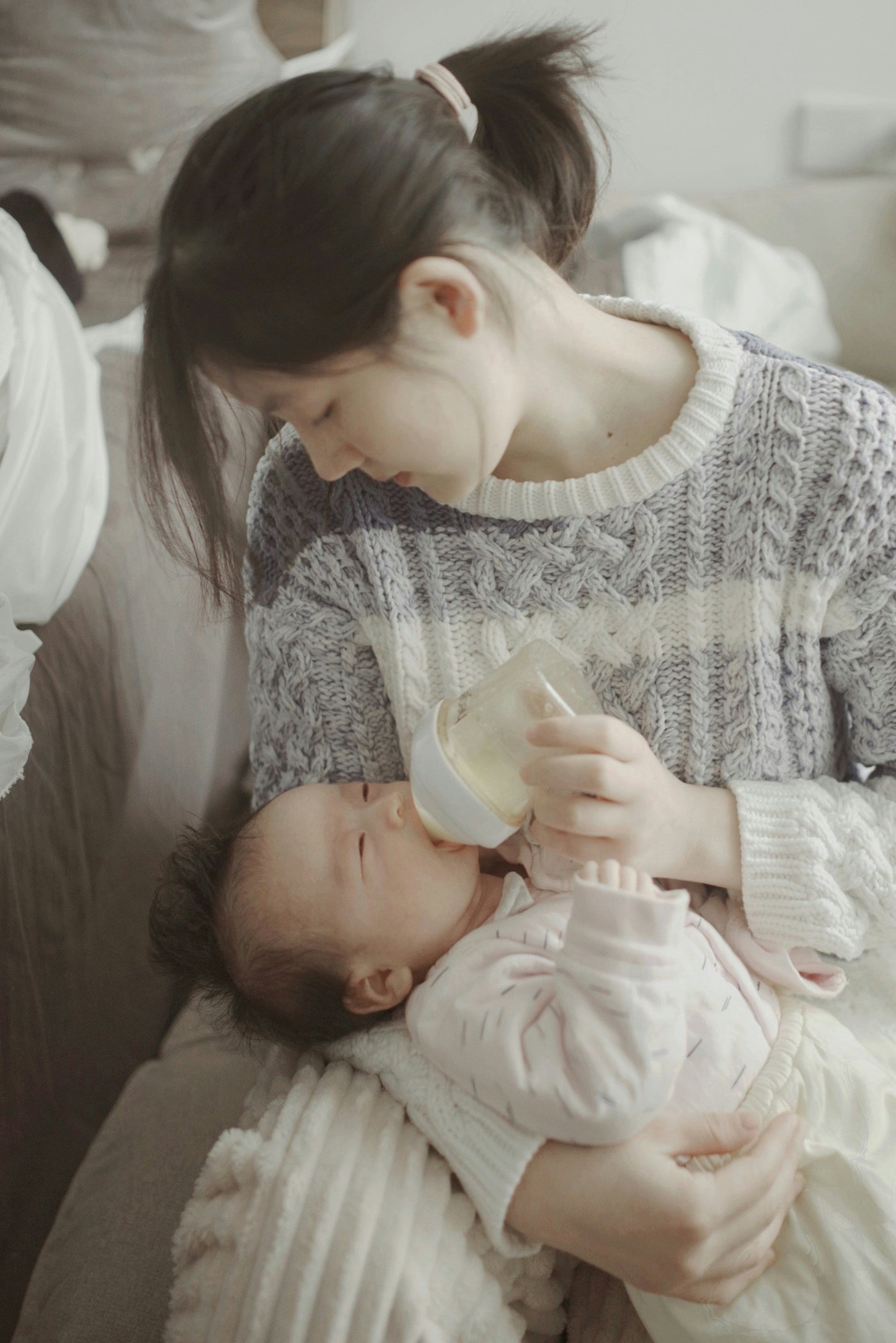 A woman in a gray sweater and ponytail looks down at her newborn, who she is feeding with a bottle.