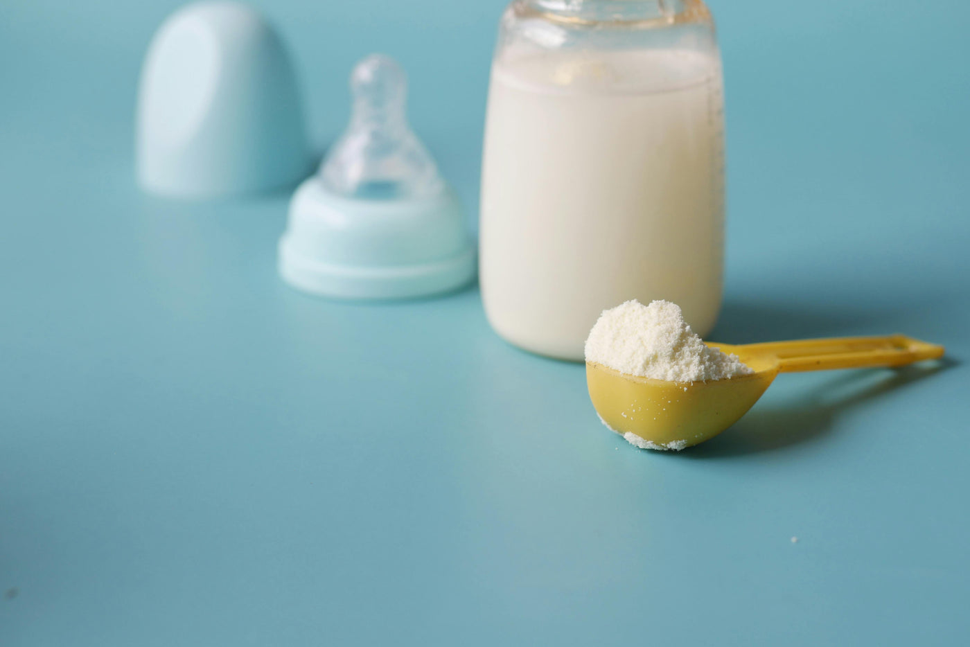 A blue background with a bottle of made-up baby formula in the back. In front, a yellow scoop of powdered baby formula sits.